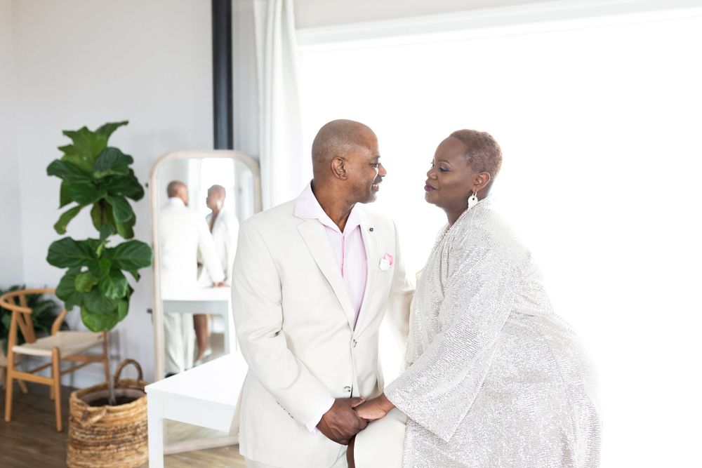 Elegant couple in white attire holding hands and gazing at each other in a bright modern room.