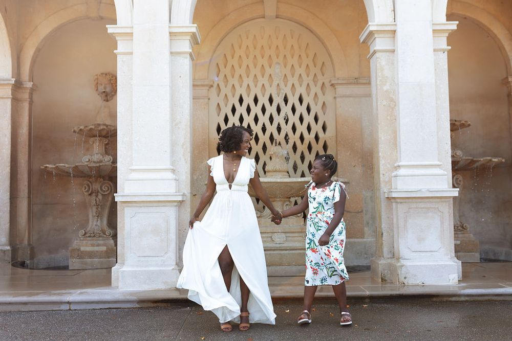 Mother in white dress holding daughters hand near ornate stone fountain in classic architecture.