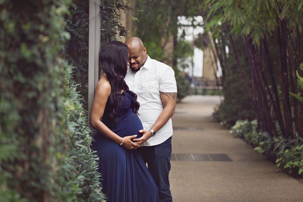 Expecting couple holding baby bump on tree-lined garden pathway.