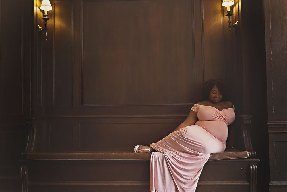 Pregnant woman in pink gown reclining gracefully on bench in elegant dark wood room.