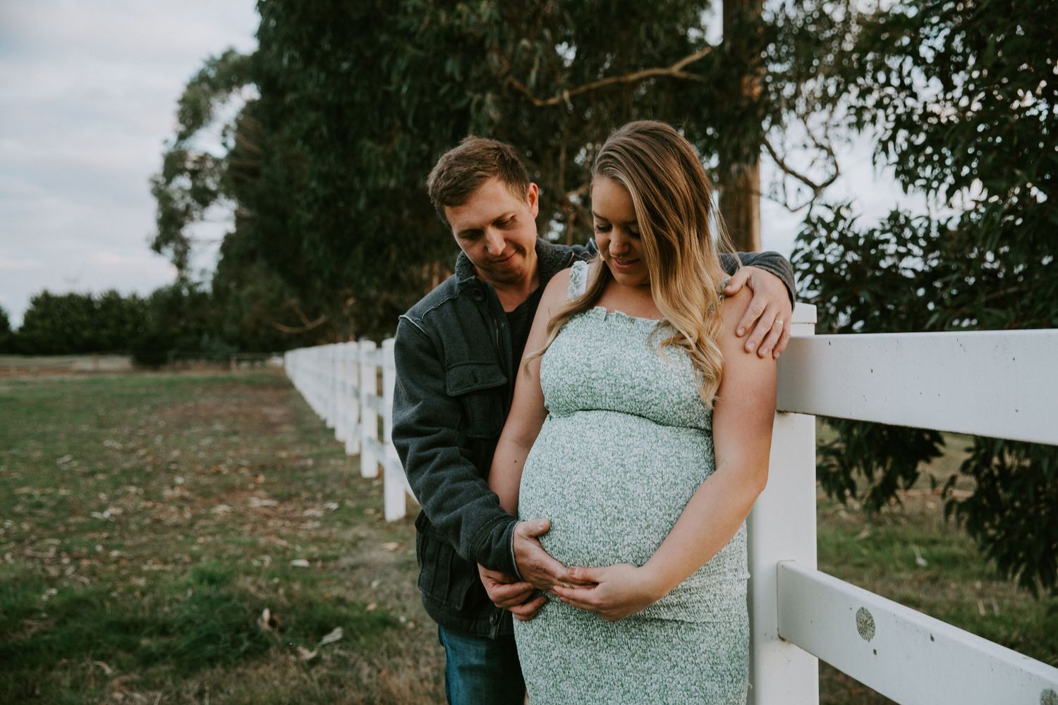 Couple embraces in a romantic outdoor setting with green trees and natural lighting in the background.