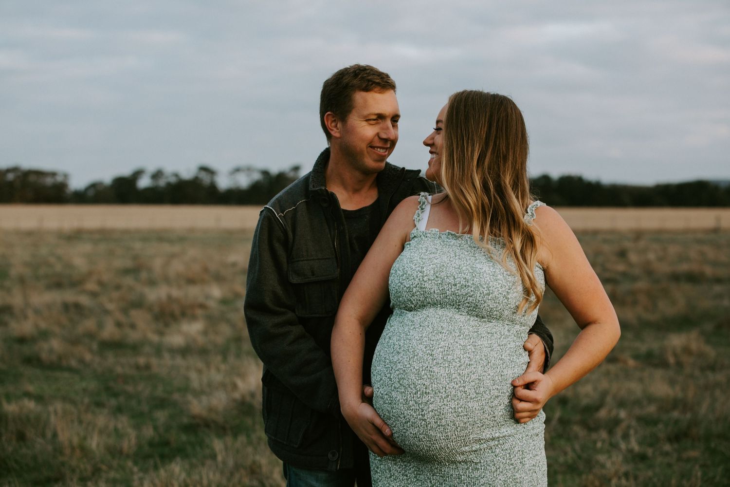 A couple embraces in a rustic field during a maternity photo session at sunset.