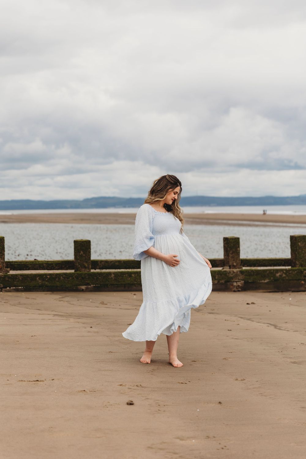 Person in white dress twirling on beach with cloudy sky and water in background.