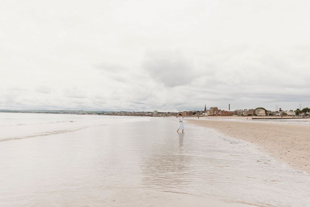 Misty coastal landscape with lone figure walking on wet sand near water's edge.