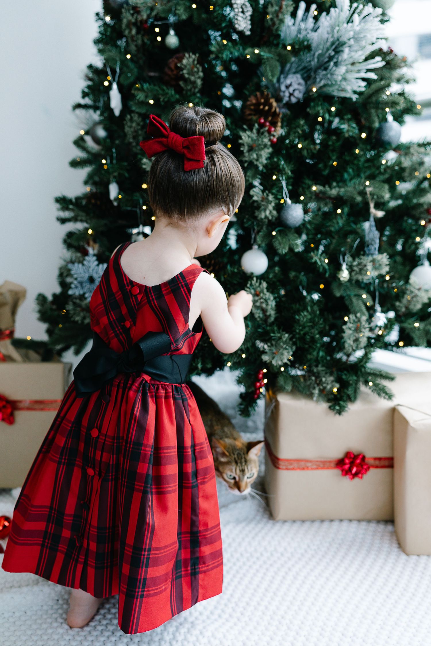 Child in red plaid holiday dress looking at decorated Christmas tree with wrapped presents nearby.