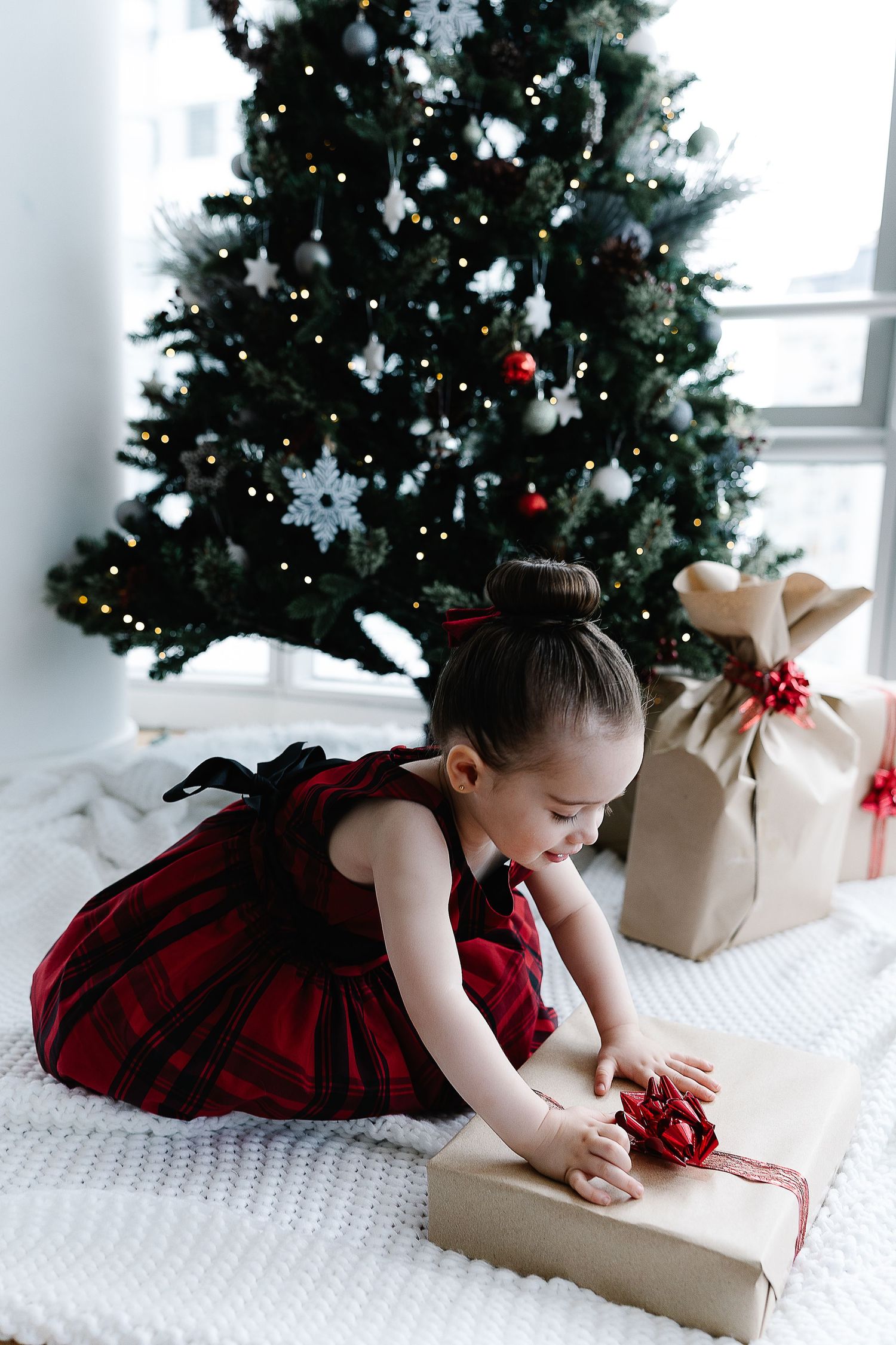 Small child in red plaid dress opening Christmas presents by decorated tree.