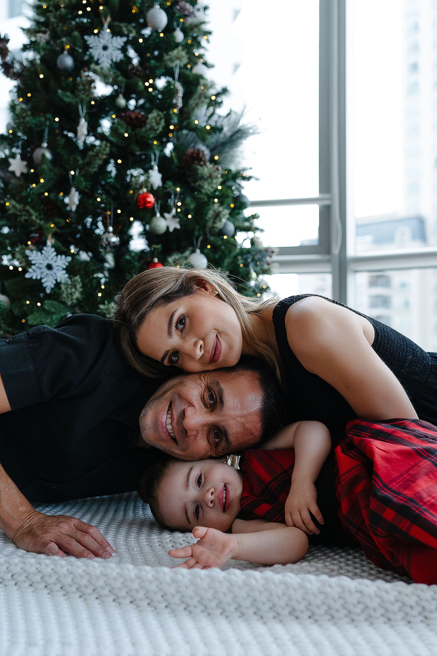Family lying together on white carpet in front of Christmas tree during holiday photoshoot.