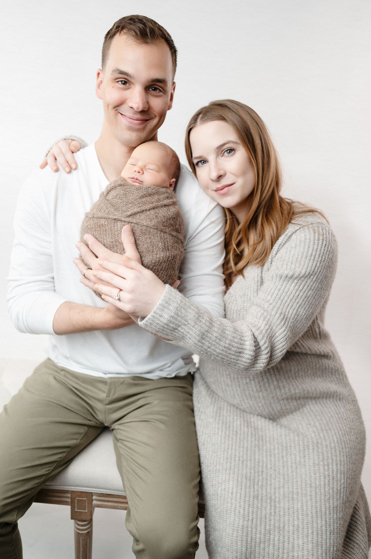 Studio family portrait with newborn baby wrapped in brown fabric against white backdrop.