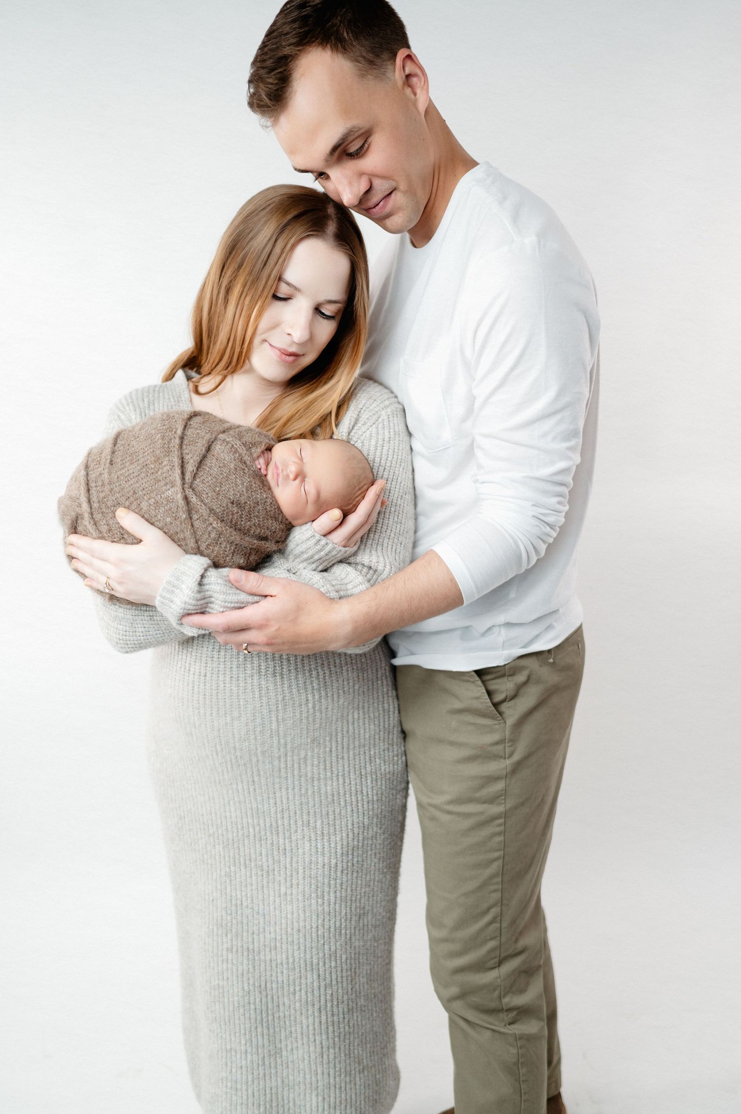 Intimate family portrait with newborn baby wrapped in brown against white studio backdrop.