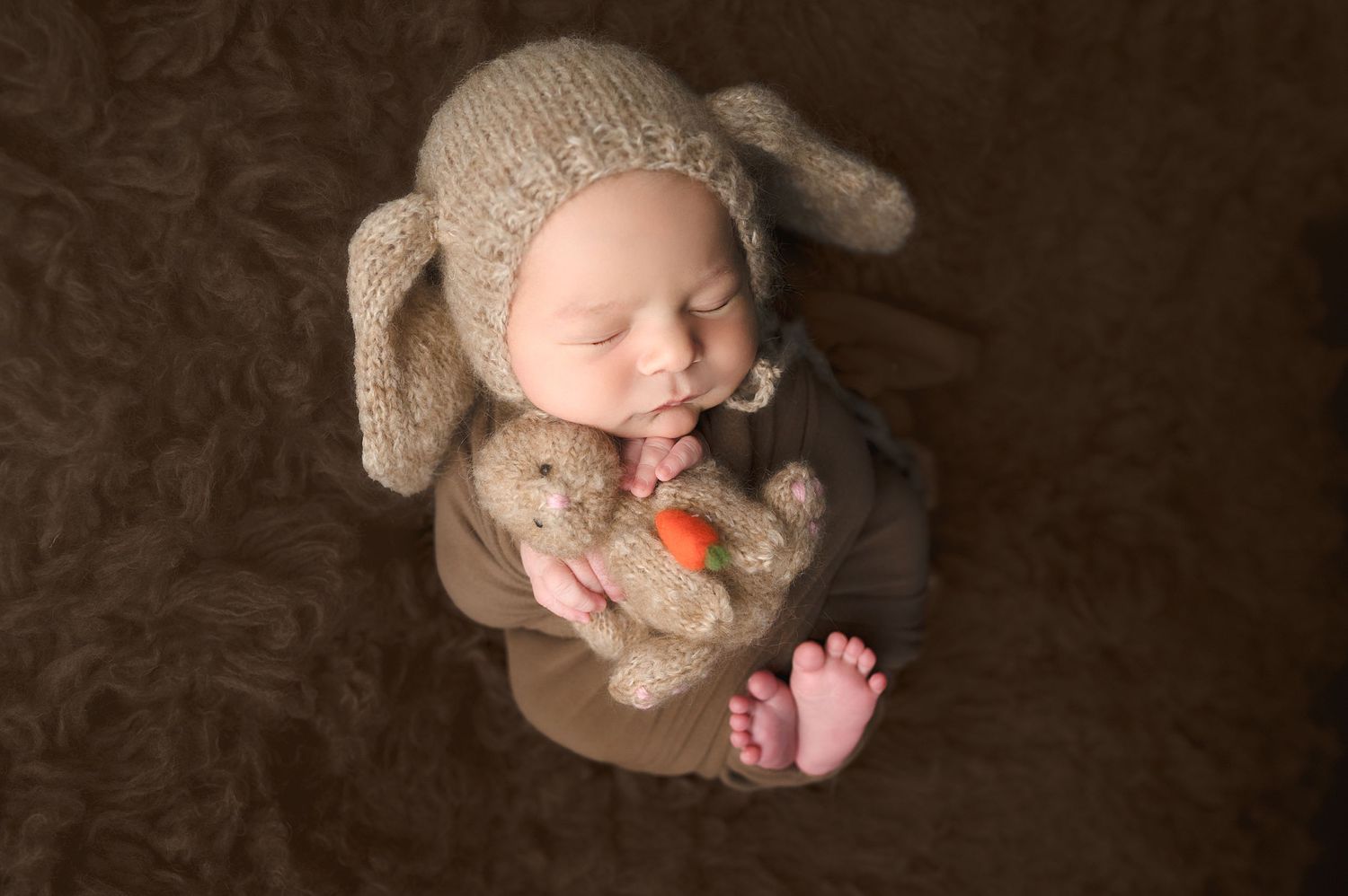 Newborn baby wearing bunny hat sleeps peacefully with stuffed toy on brown textured backdrop.