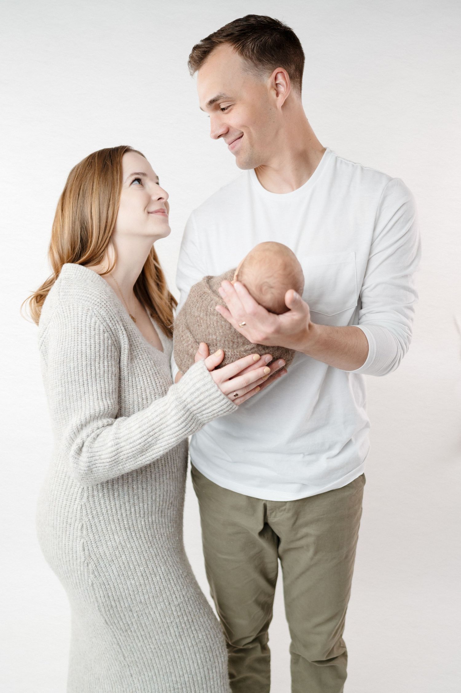 Loving parents share tender moment while holding their swaddled newborn against white background.