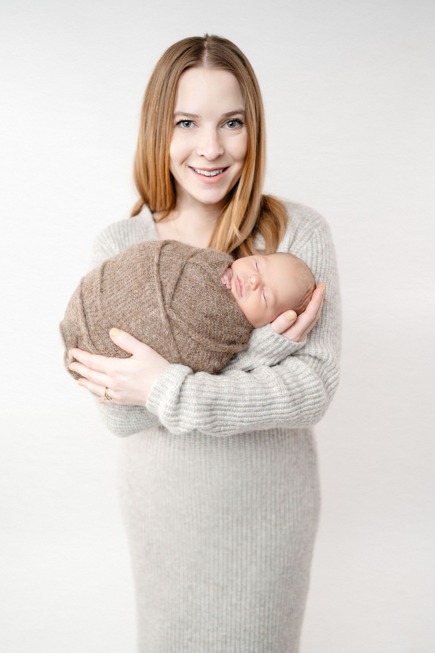 Parent in gray sweater dress holding sleeping newborn wrapped in brown blanket.