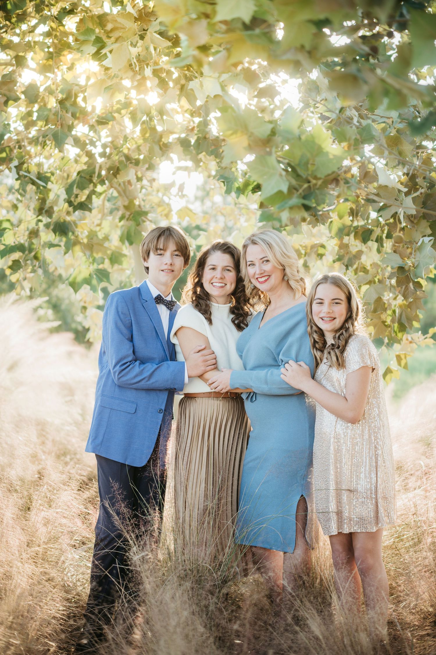 A family photo captured under sunlit tree branches in a dreamy outdoor setting.