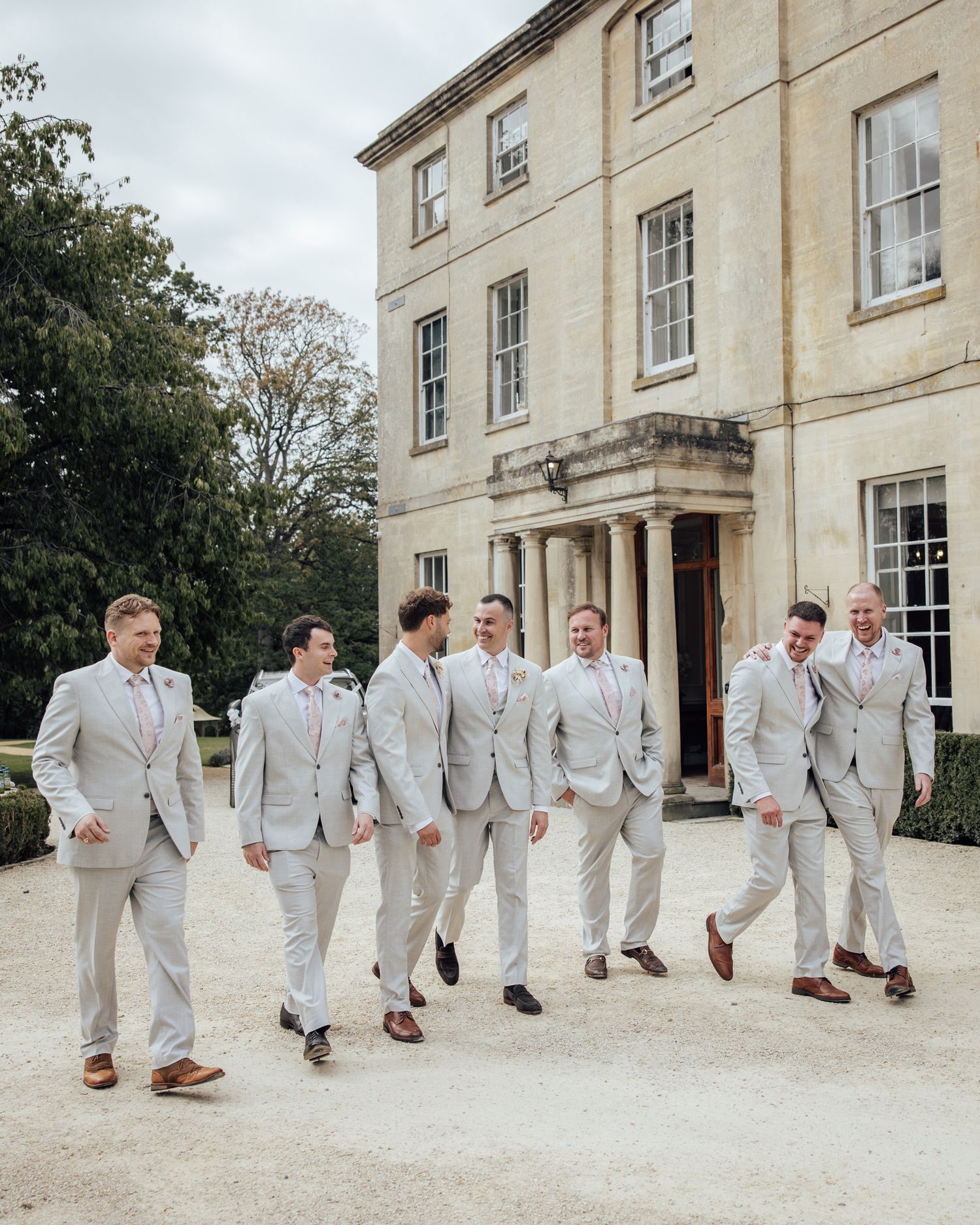 Groomsmen in light gray suits walking together outside a grand stone manor house on a wedding day.
