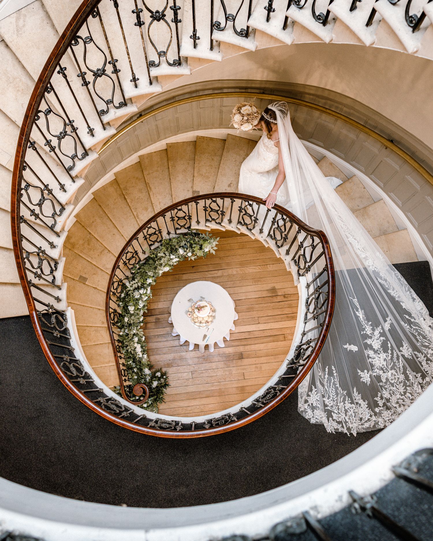 Aerial view of a bride in a white gown and lace veil descending an elegant spiral staircase adorned with greenery.