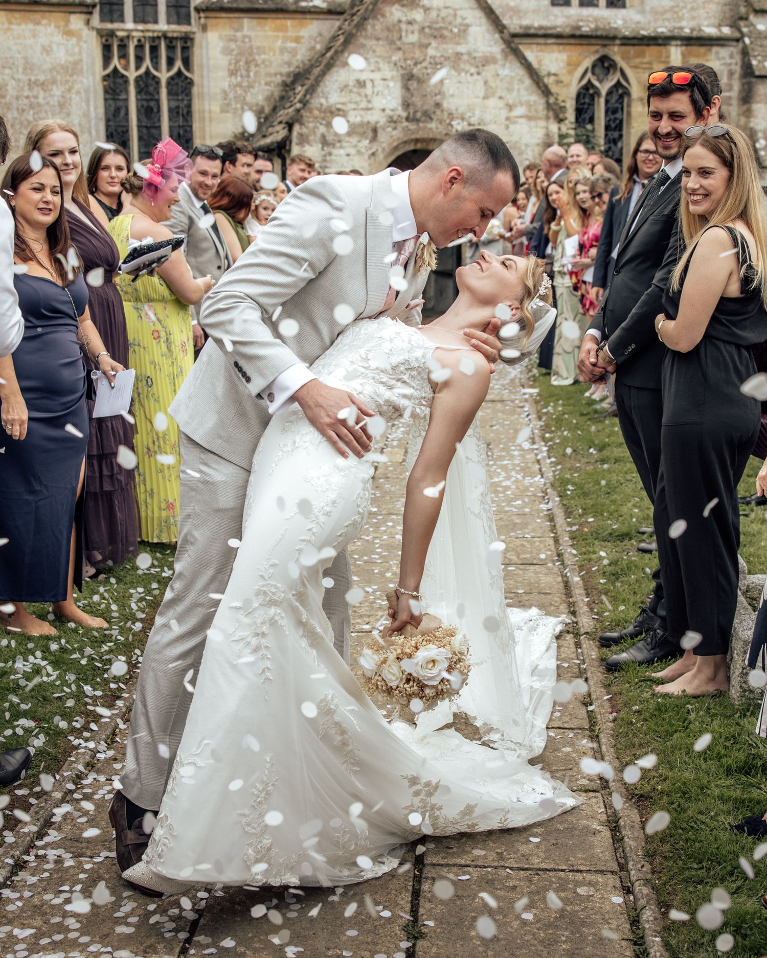 Bride and groom share a romantic dip kiss surrounded by white confetti outside a historic church.