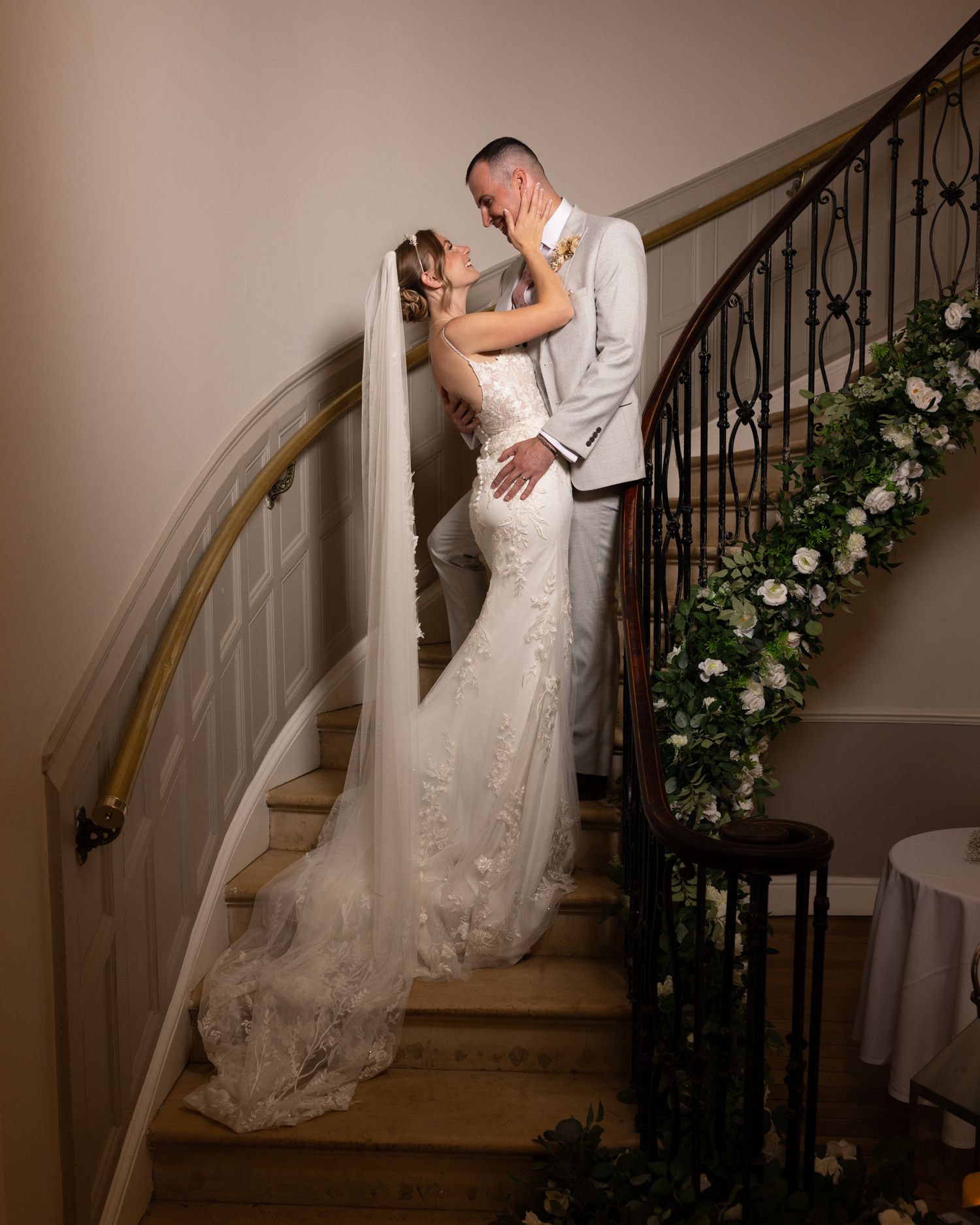 Bride and groom share a romantic kiss on an elegant staircase adorned with lush greenery and white roses.