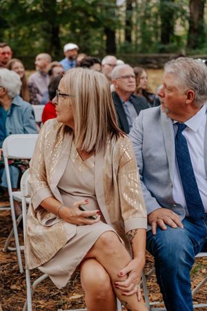 Elegantly dressed wedding guests seated outdoors, woman in gold sequin jacket and man in grey suit with blue tie.