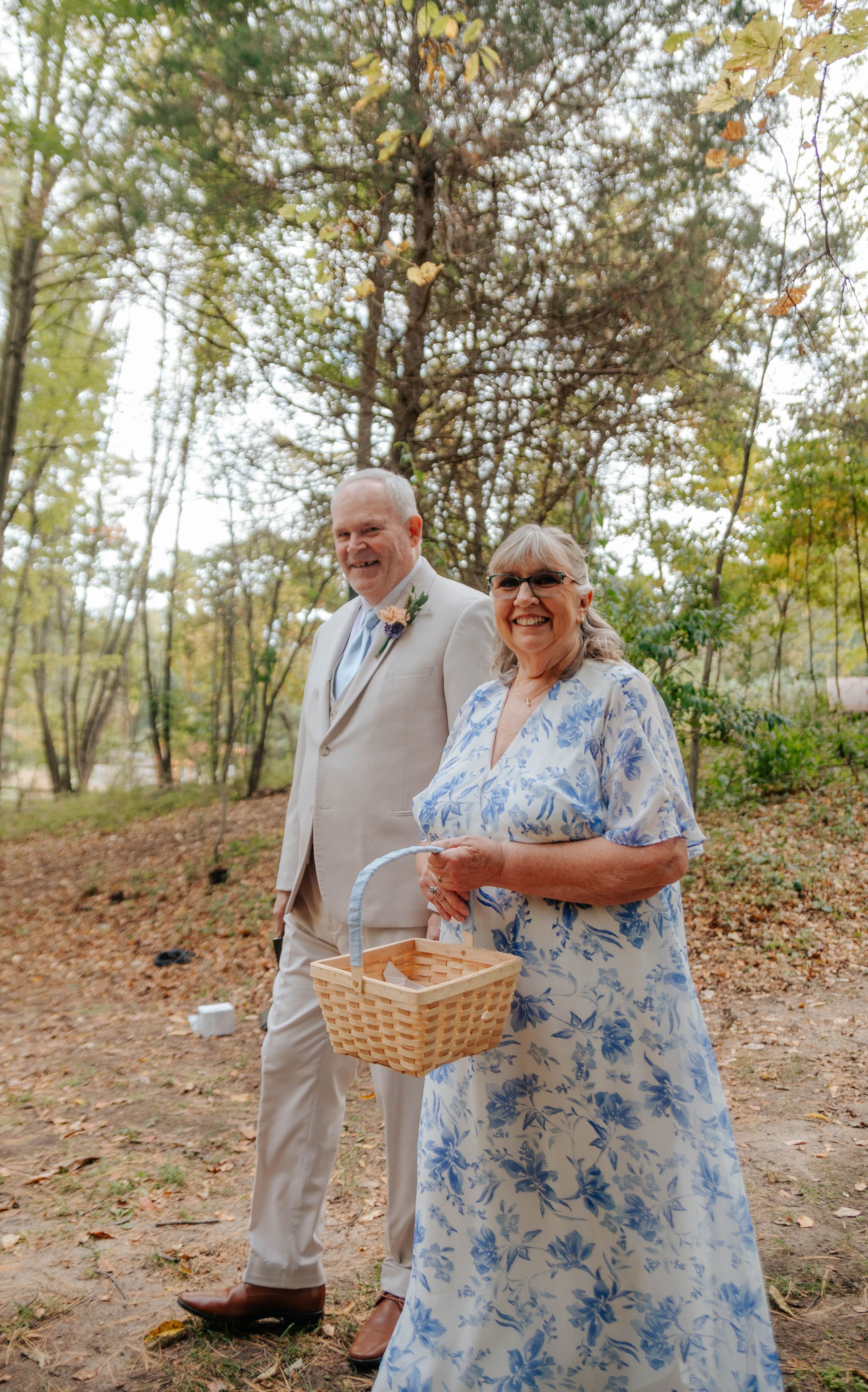 Elderly couple smiling outdoors at a wedding, man in tan suit, woman in blue floral dress holding a wicker basket.