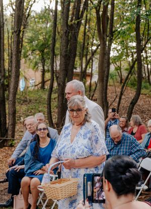 Elderly woman in blue floral dress carrying basket walks down outdoor woodland wedding aisle.