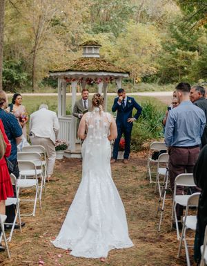Bride in lace gown walks down outdoor aisle toward gazebo as groom wipes tears at fall wedding ceremony.