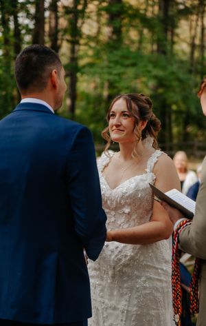 Bride in lace wedding dress smiling at groom in navy suit during outdoor forest wedding ceremony.