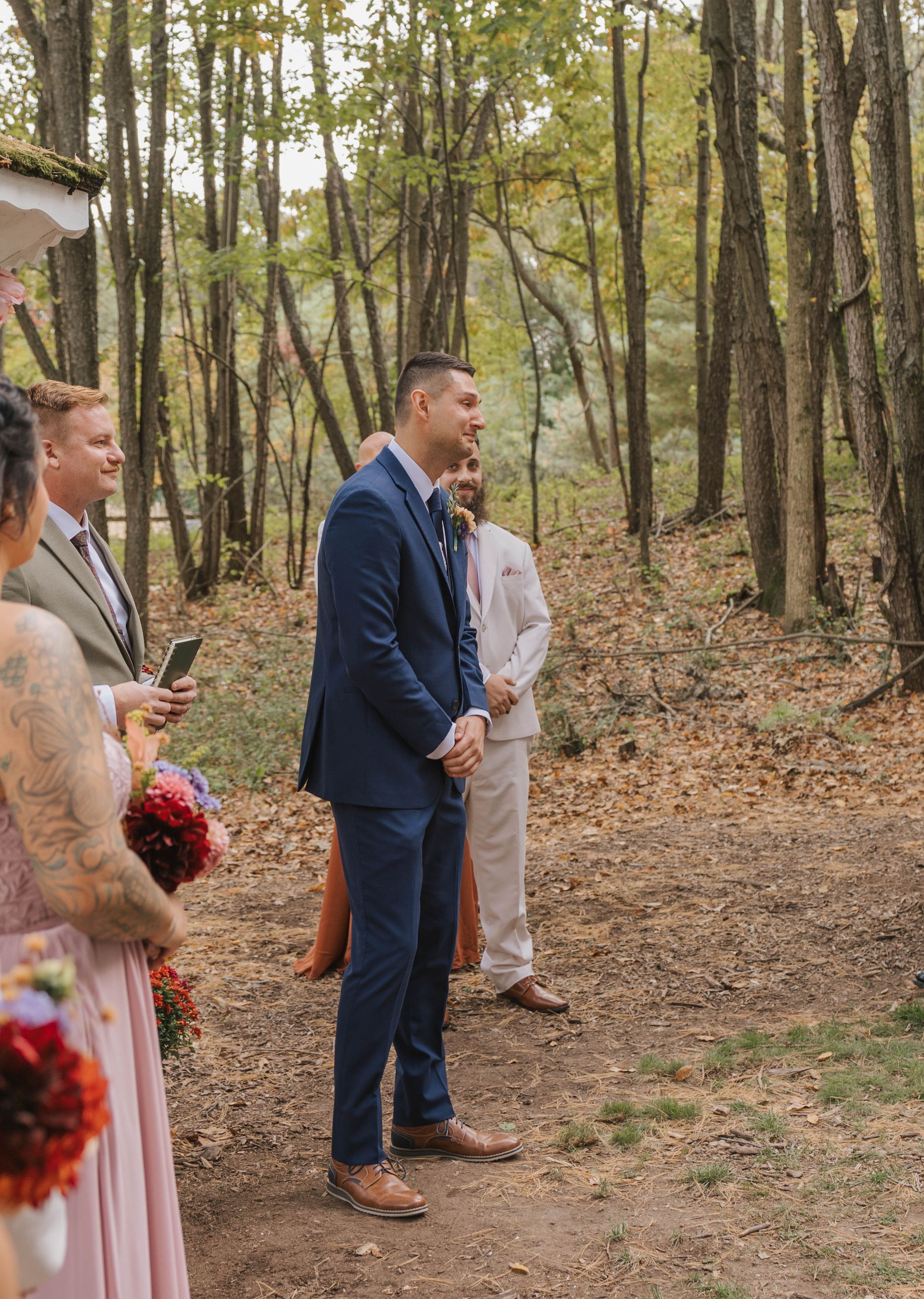 Groom in navy blue suit standing at outdoor woodland wedding ceremony with wedding party on forest path.
