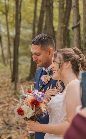 A smiling bride holding a colorful fall bouquet stands beside her groom in a blue suit during an outdoor autumn wedding ceremony.