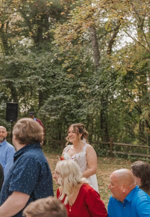 Smiling bride in white floral dress laughing with wedding guests outdoors among autumn trees.