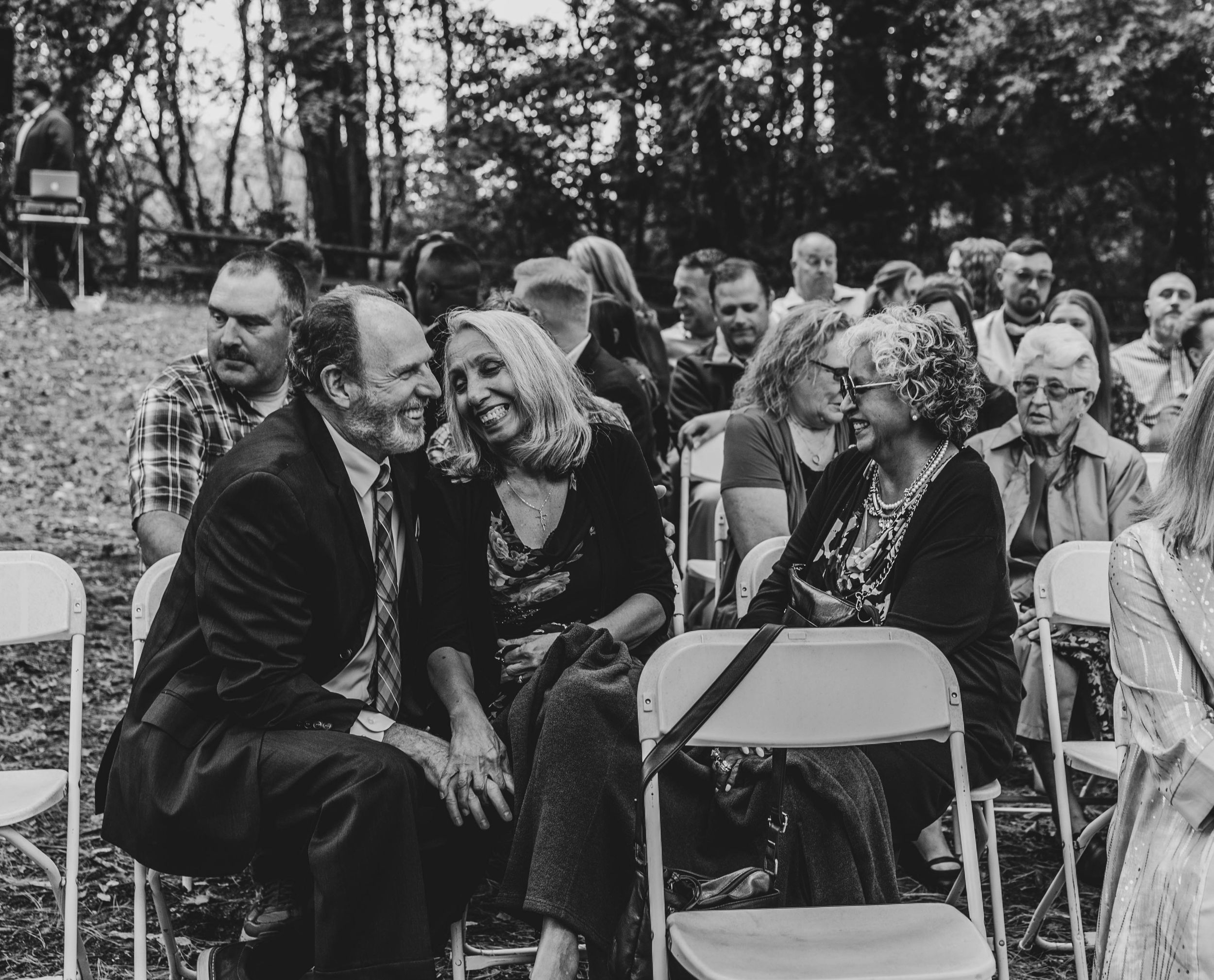 Black and white outdoor wedding ceremony guests smiling and laughing while seated on white folding chairs.