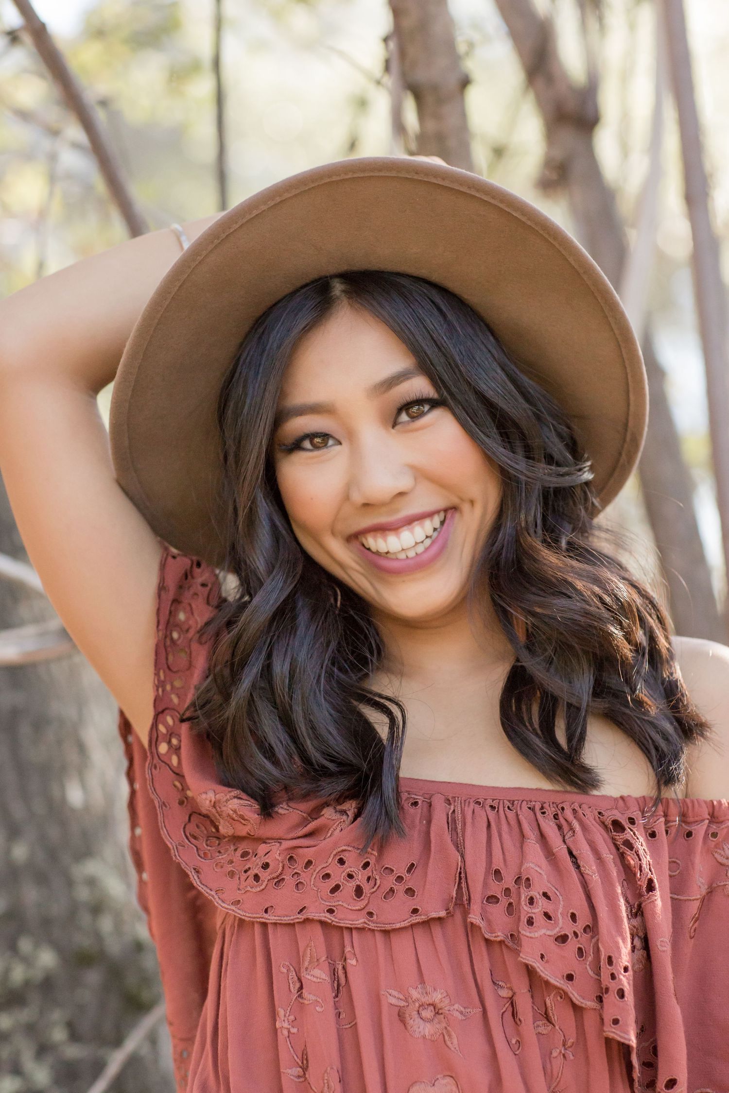 Smiling young woman in rust off-shoulder top wearing a tan wide-brim hat outdoors among trees.