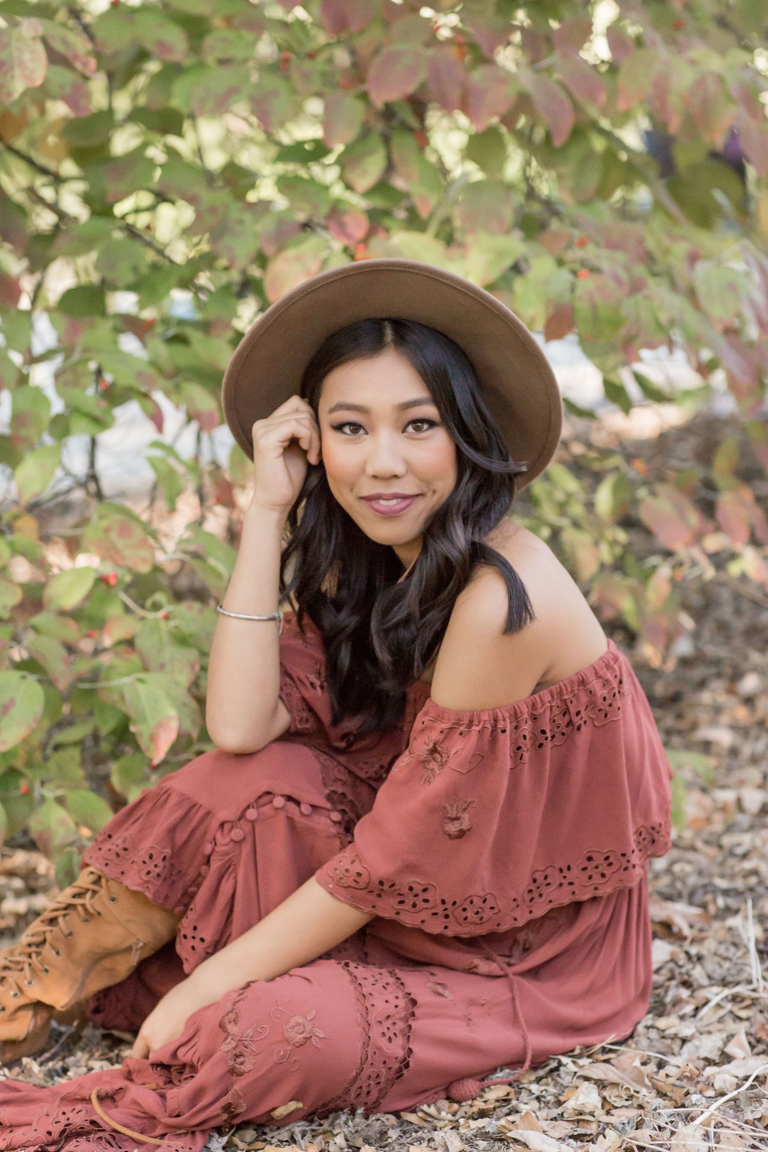 Young woman in rust boho off-shoulder dress and tan hat sitting outdoors among colorful fall foliage.
