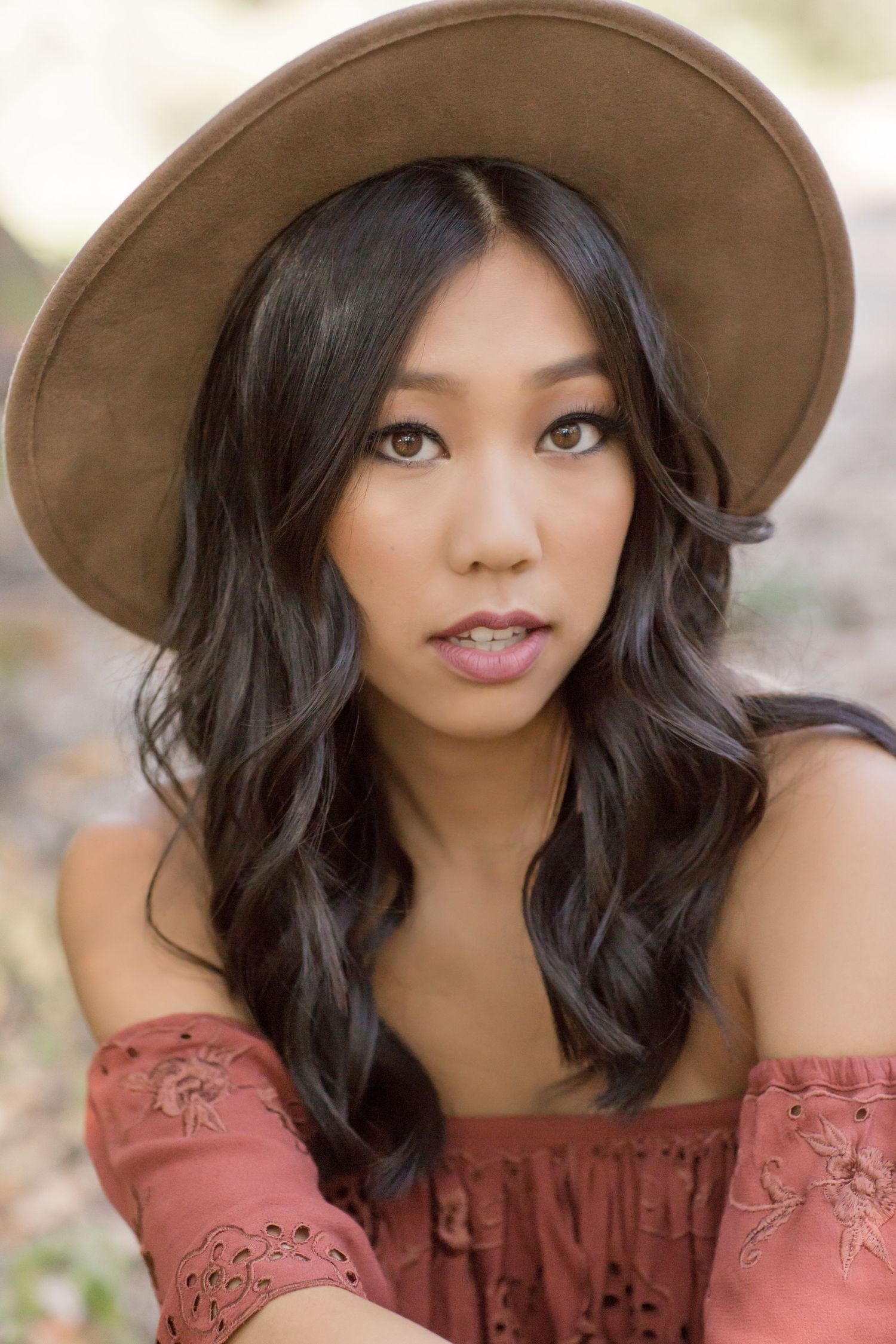 Close-up of young Asian woman in rust off-shoulder top wearing a tan wide-brim hat outdoors.