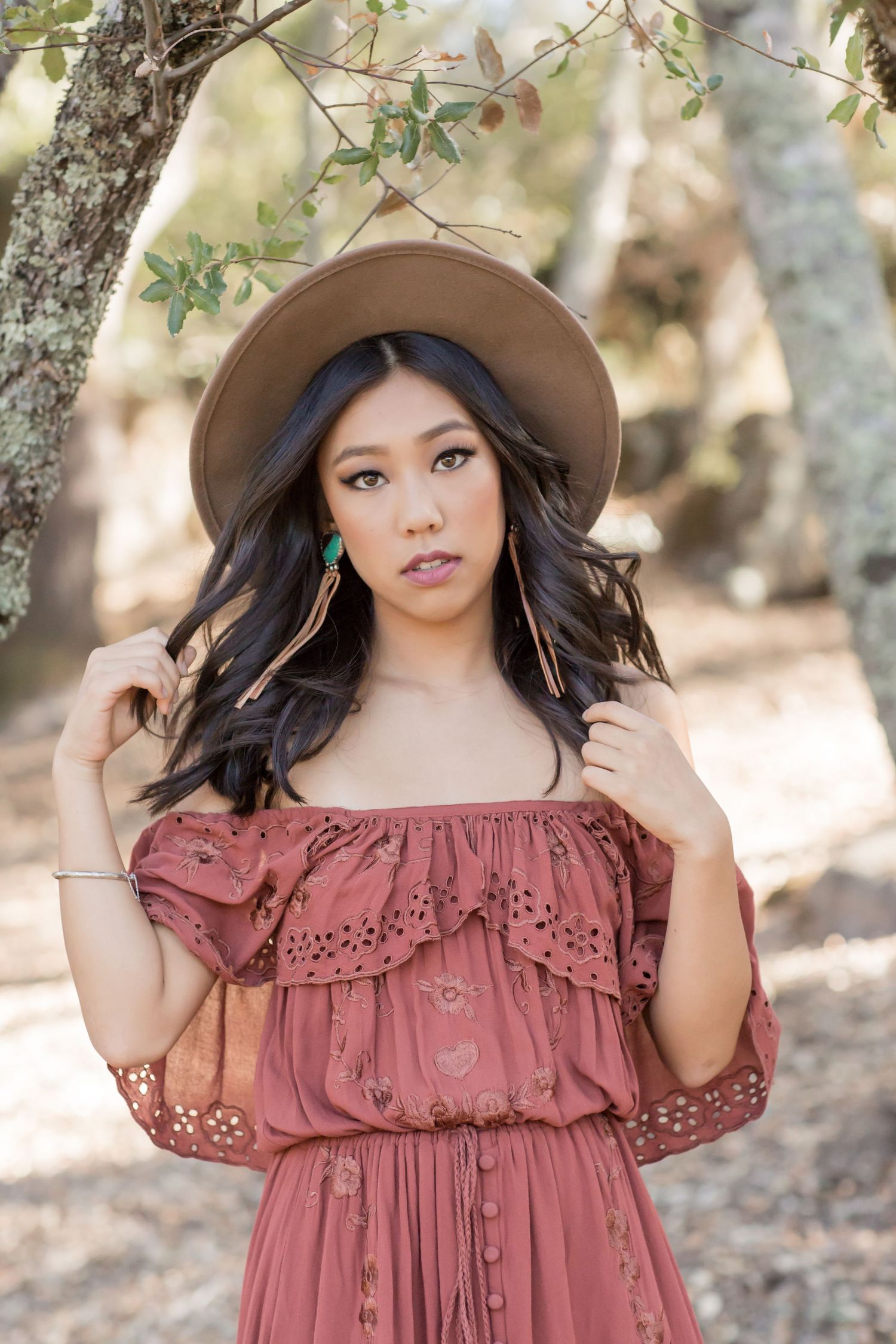 Young Asian woman in rust boho maxi dress and tan hat posing among trees in natural setting.