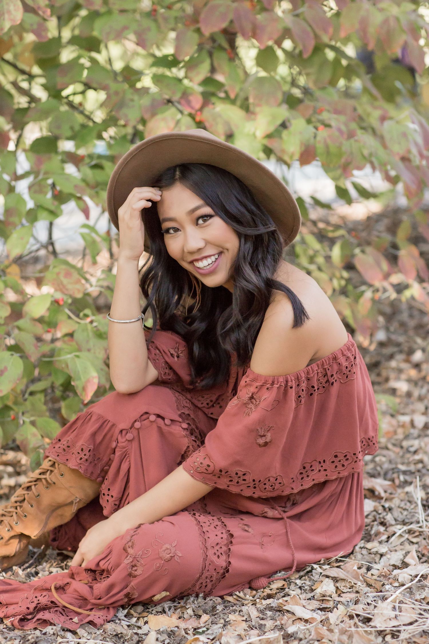 Smiling young woman in rust off-shoulder boho dress and tan hat sitting among fall foliage outdoors.