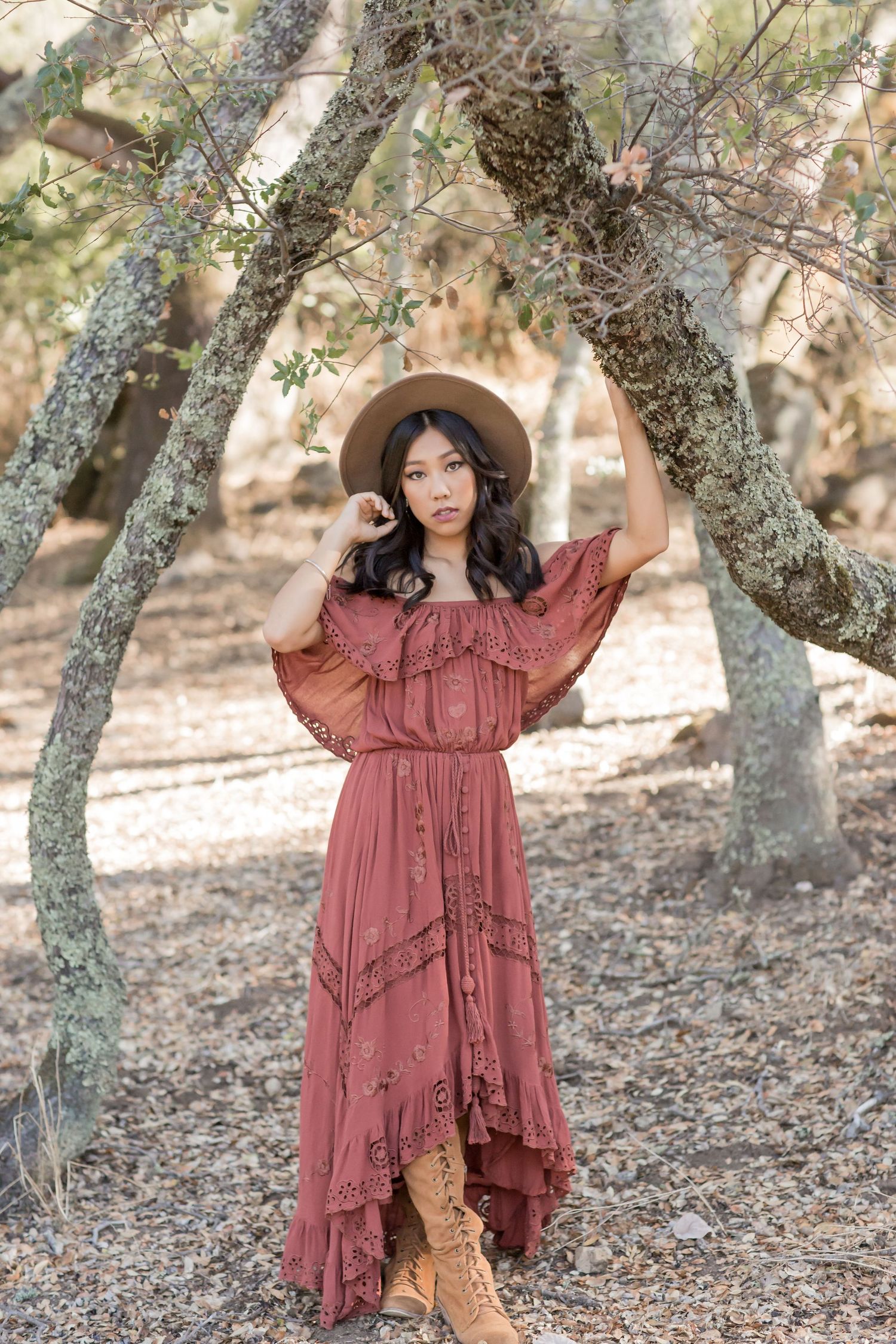 Young woman in rust boho maxi dress and tan hat posing under arched tree branches outdoors.