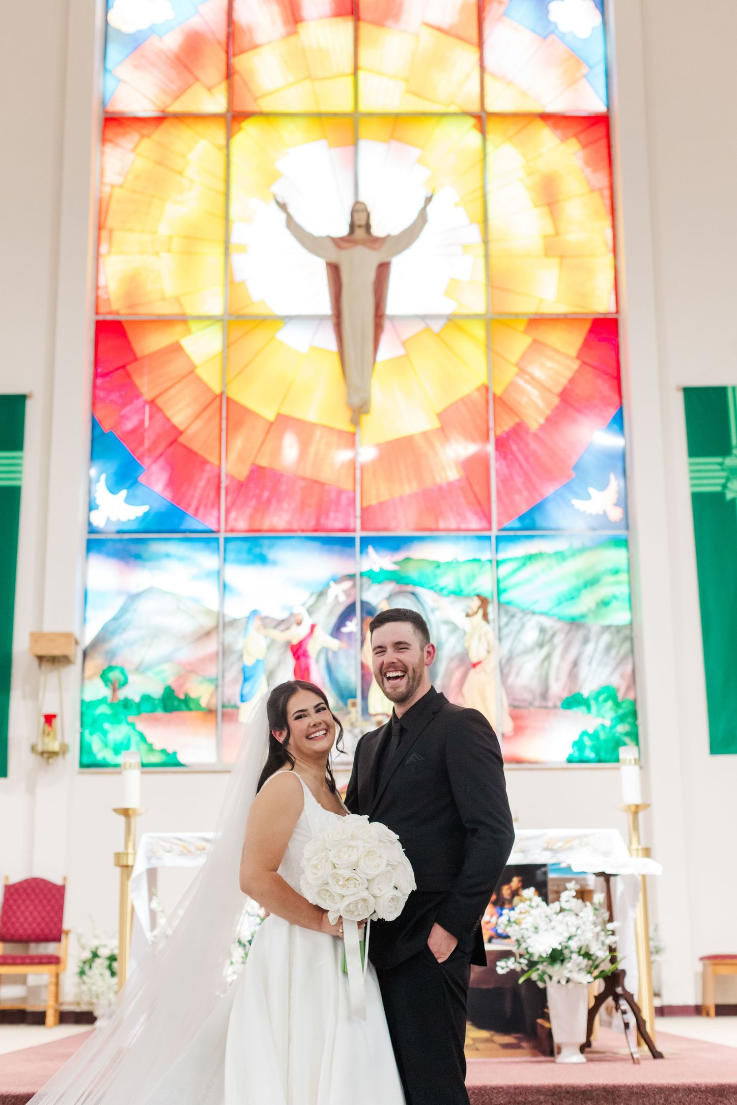 Smiling bride and groom pose joyfully before a colorful stained glass window with a Christ figure inside a church, holding a white bouquet.