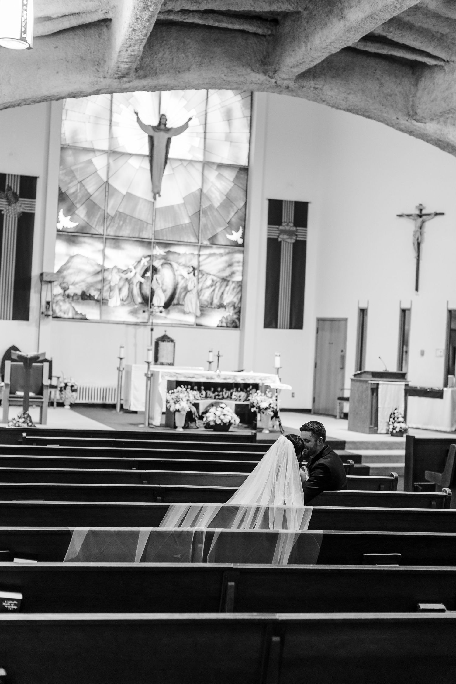Black and white photo of a bride with a long veil seated in a church pew facing the altar, with a crucifix and stained glass Christ window.