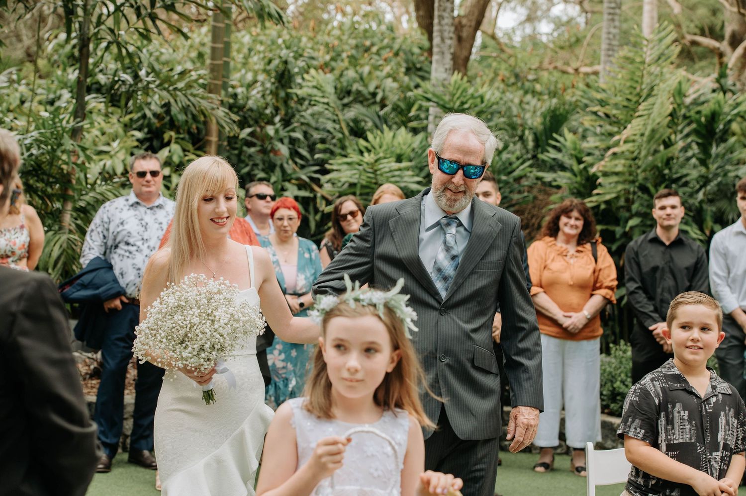 Wedding party walking down a lush garden path with bride in white dress carrying baby's breath bouquet.