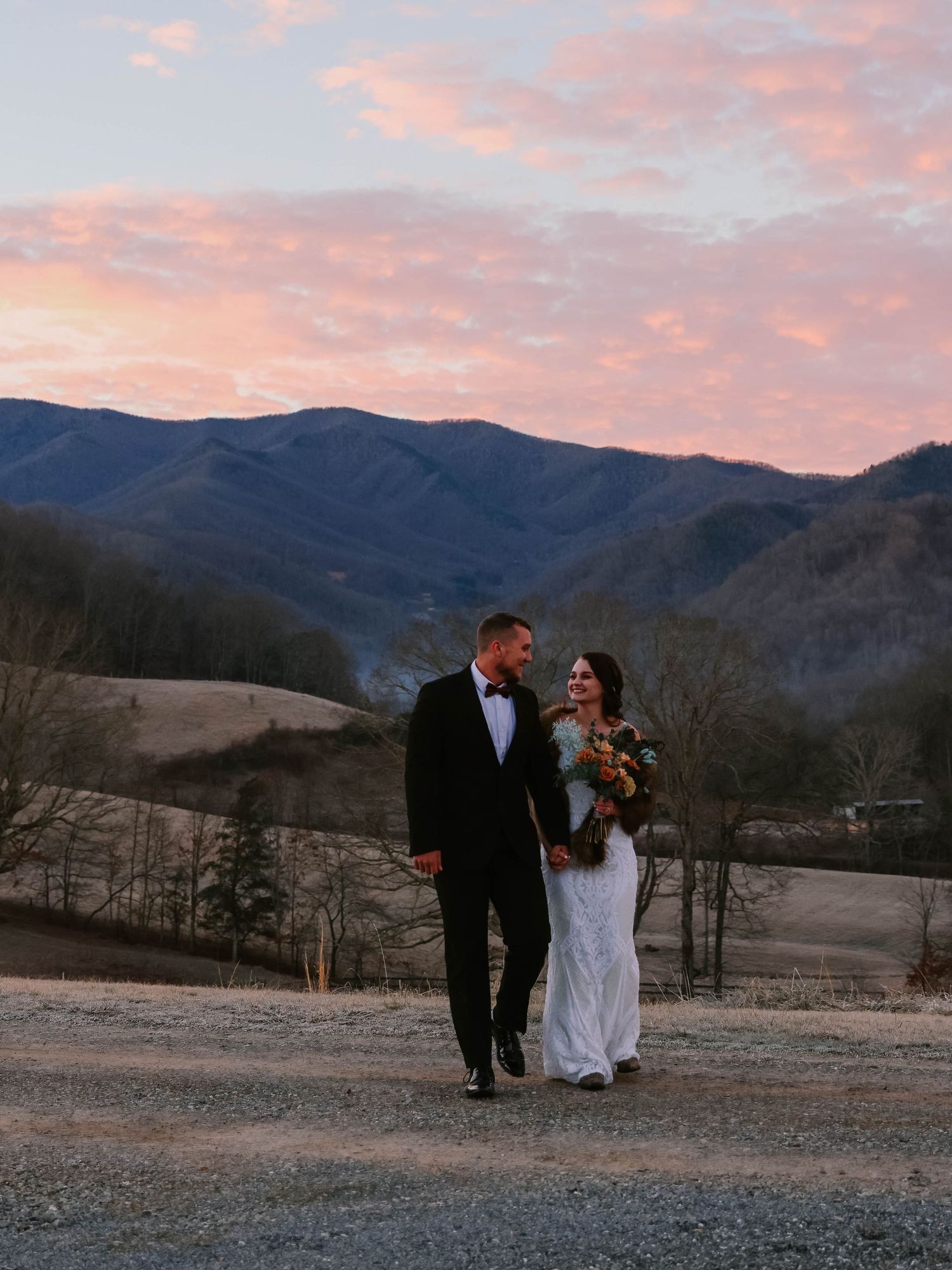 Romantic mountain sunset wedding photo with couple on dirt road against pink and purple sky.