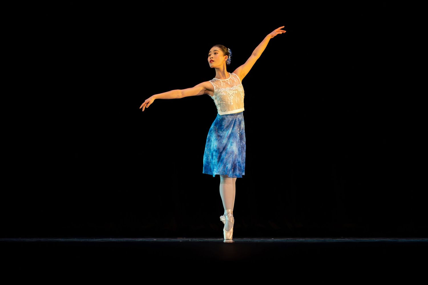 Ballet dancer in blue skirt performs graceful balance pose en pointe against dark background.