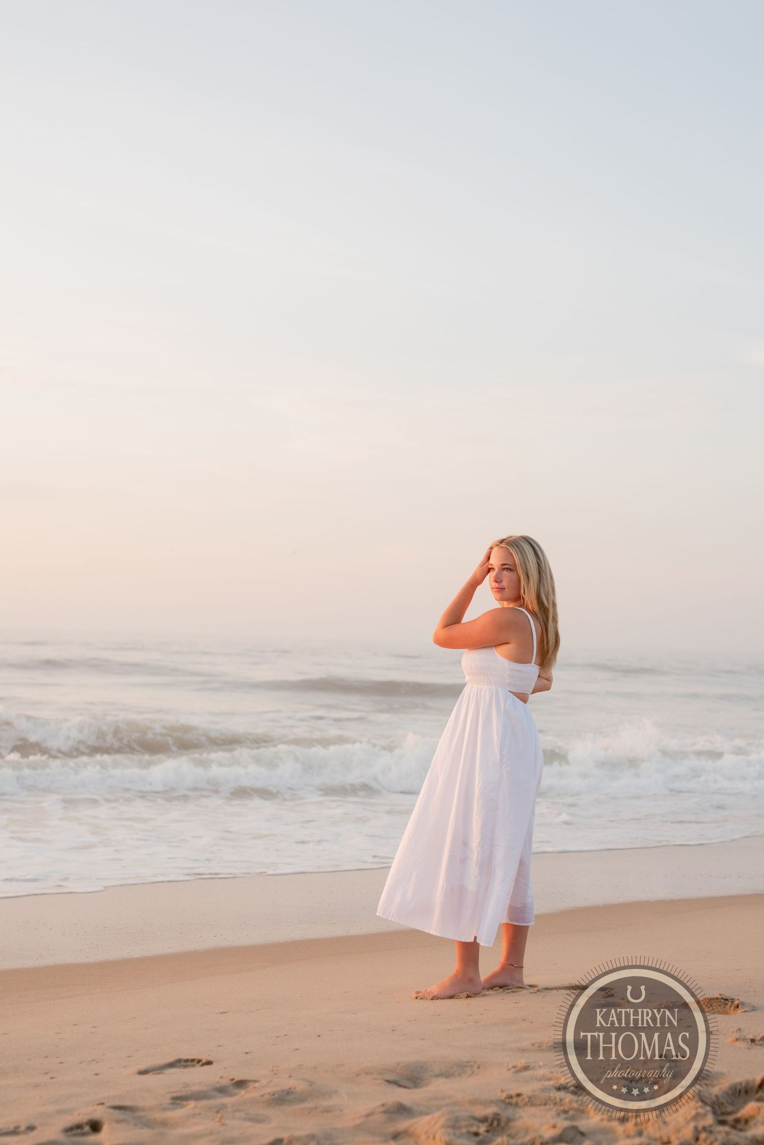 Someone in a white dress stands on a beach with crashing waves and soft pastel sky.