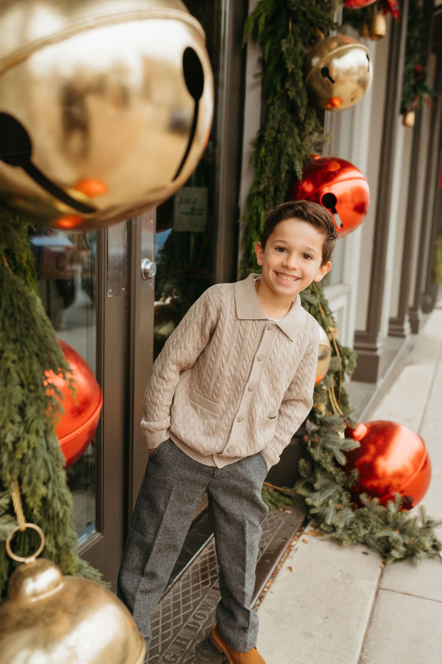 A smiling boy in a beige cable-knit sweater and gray pants poses outdoors among festive Christmas ornaments and bells.