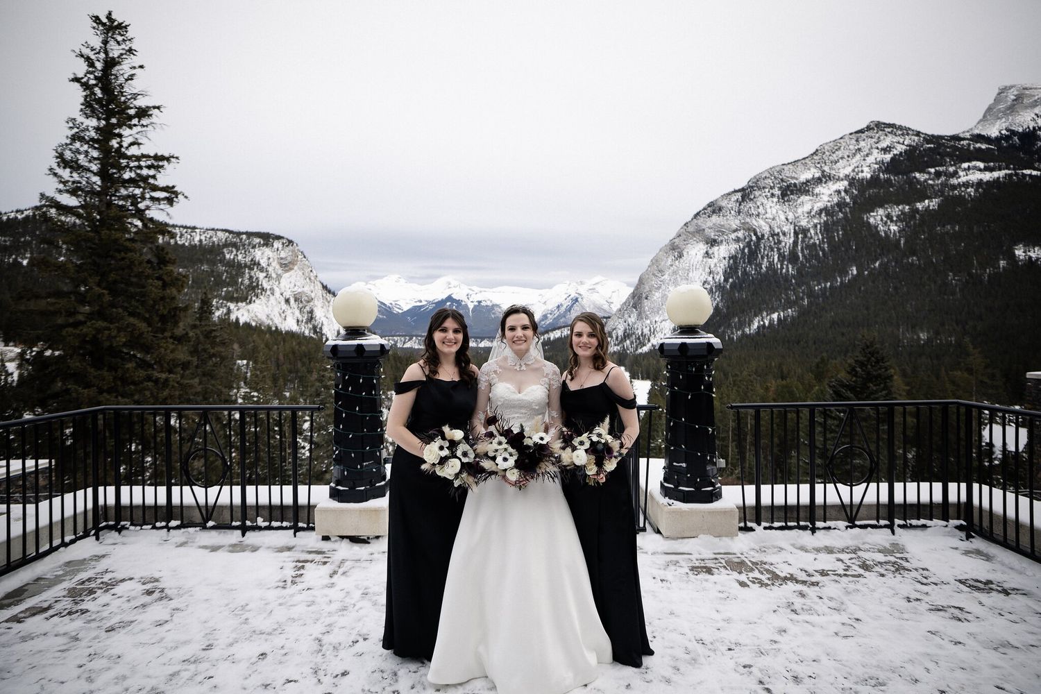 Bride in a lace gown with two bridesmaids in black on a snowy terrace with mountains at a Banff winter wedding