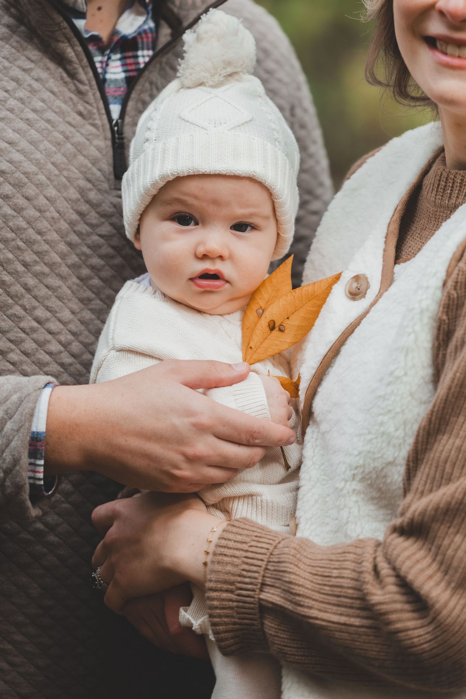A baby in a white knit hat and vest holds an autumn leaf while being cradled by parents outdoors in fall in Lake Fairfax