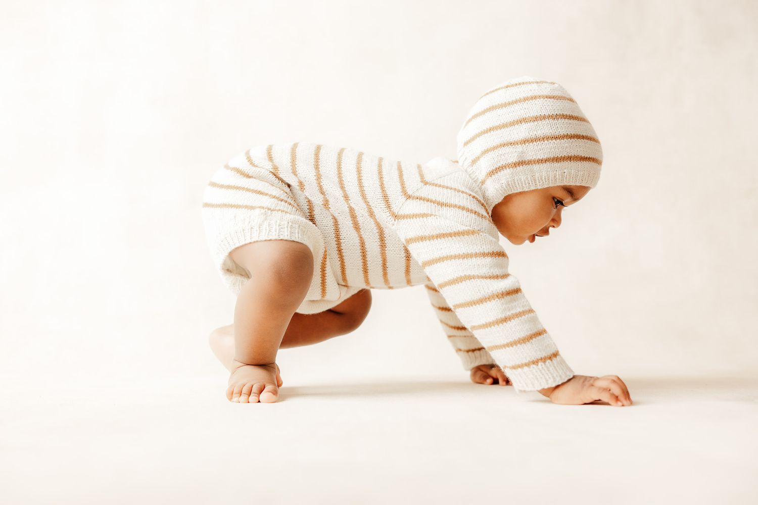 Baby in striped knit romper and hat crawling on white floor, side profile view.