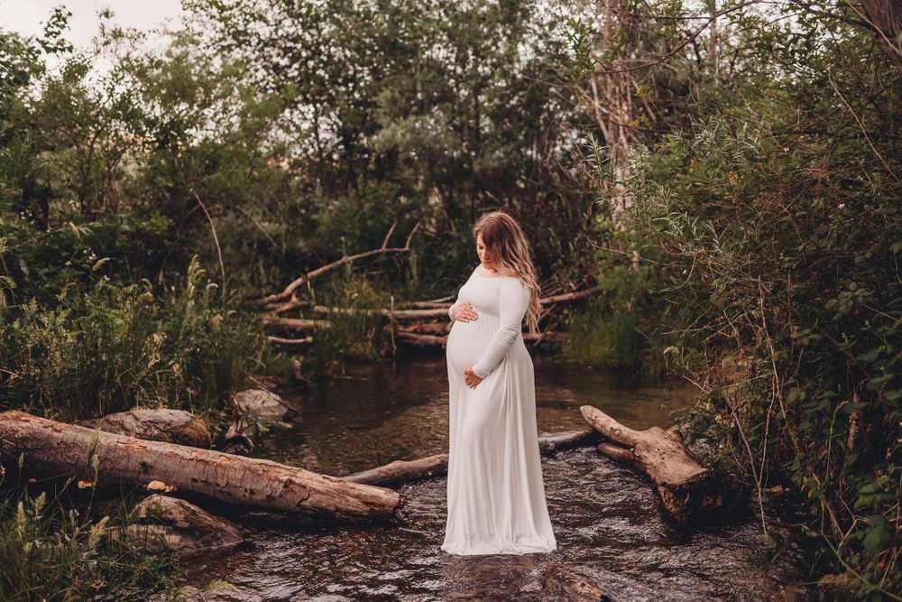 Maternity photo of woman in white dress standing on fallen logs over stream.