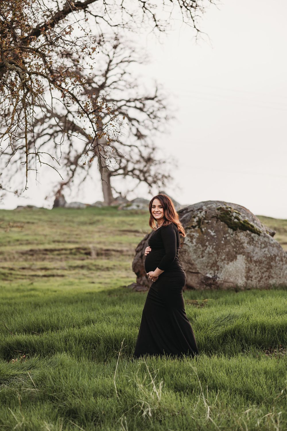Maternity photo of woman in black dress against hillside with large boulder.