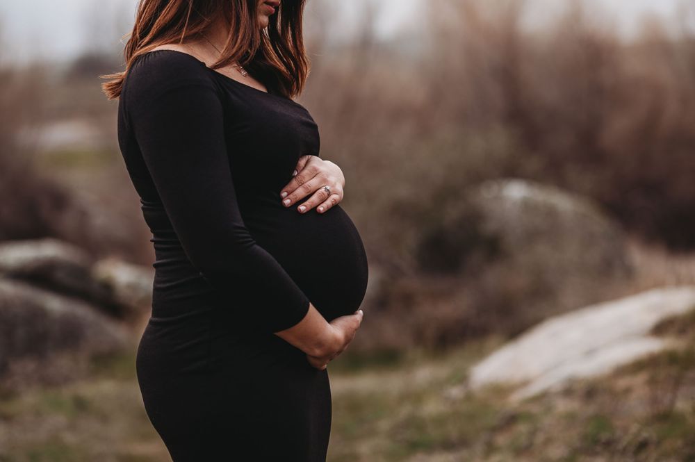 Side view of pregnant belly in fitted black dress against blurred natural background.