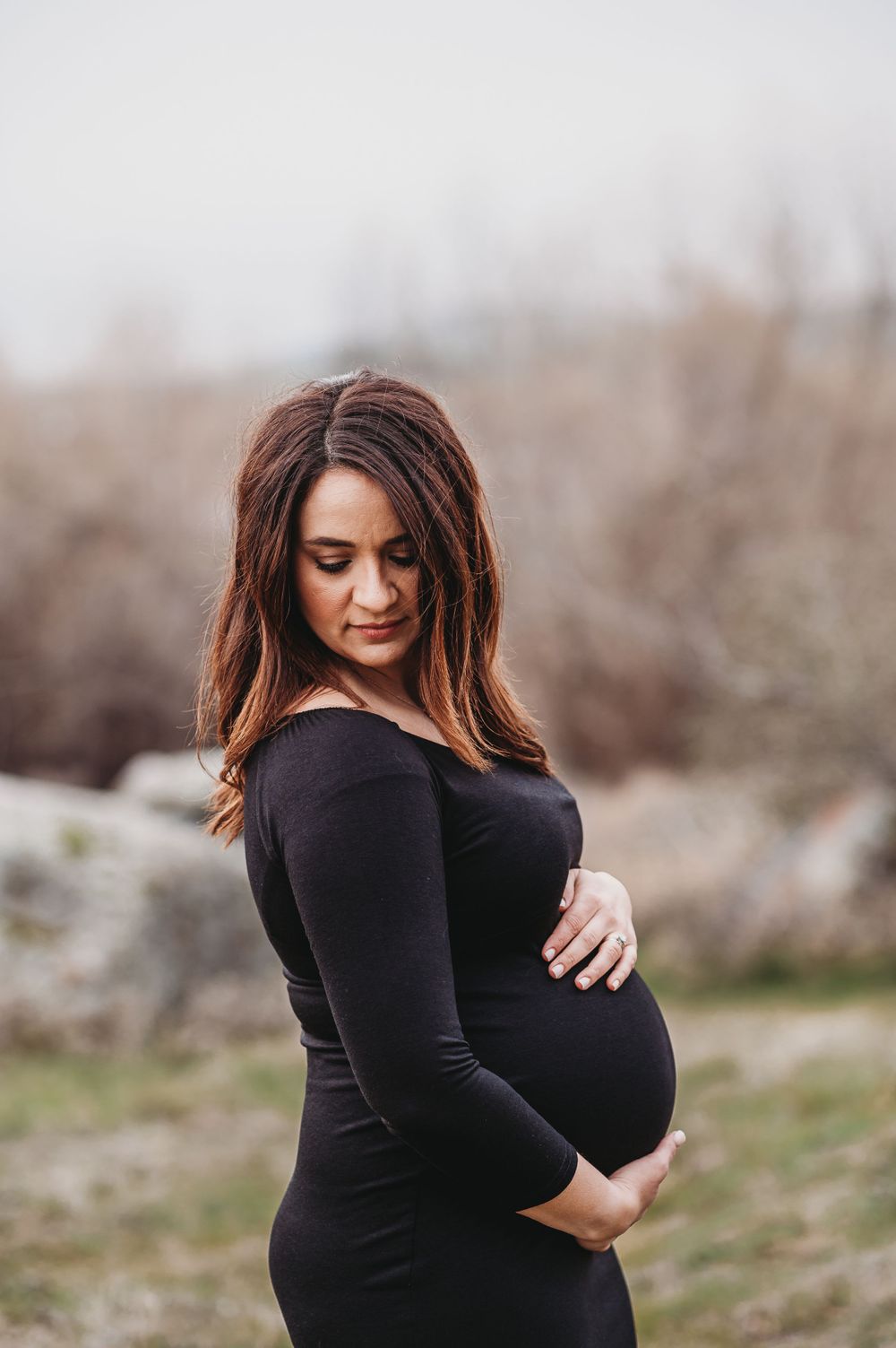 Elegant maternity portrait featuring black form-fitting dress against natural backdrop.